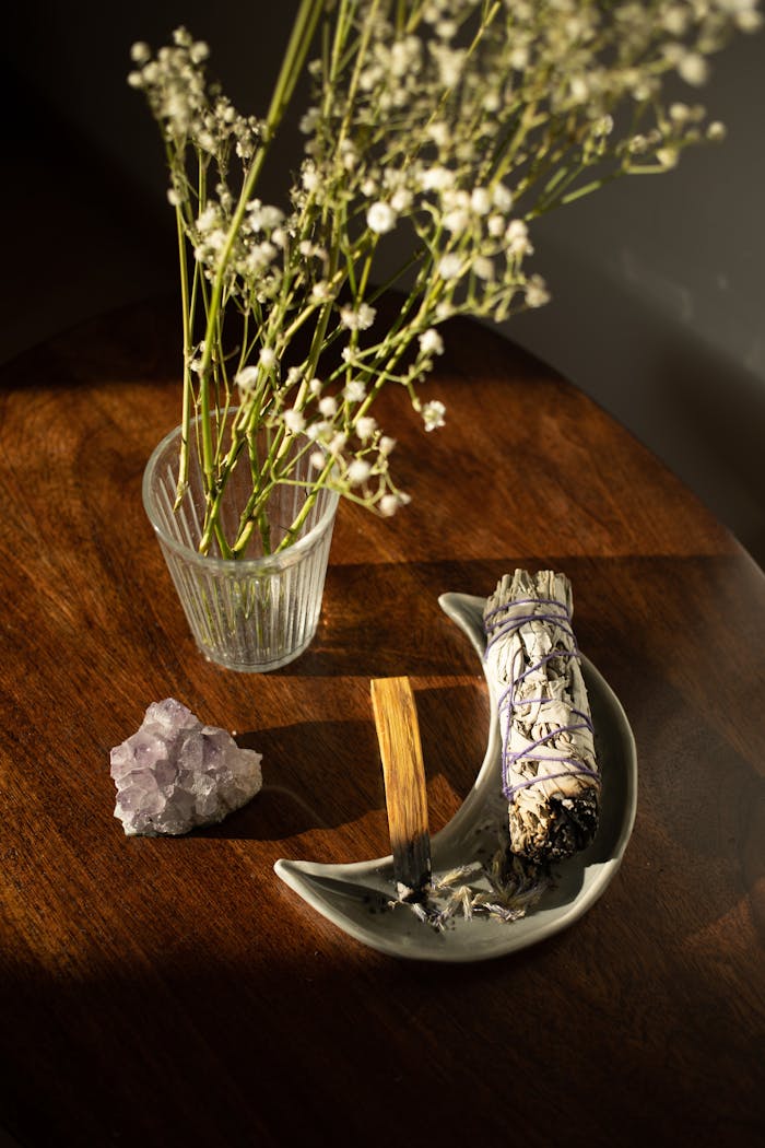 Still life of white sage bundle, amethyst, flowers, and incense on a wooden table.